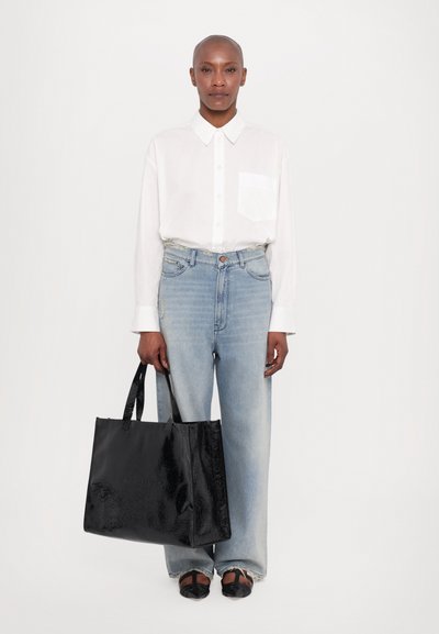 Black patent tote bag with two handles. The bag features a structured shape and shiny texture, contrasting with light blue wide-leg jeans and a white shirt.