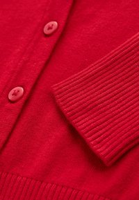 Cardigan rouge en tricot montrant un poignet côtelé, une patte de boutonnage et des boutons rouges assortis en gros plan.