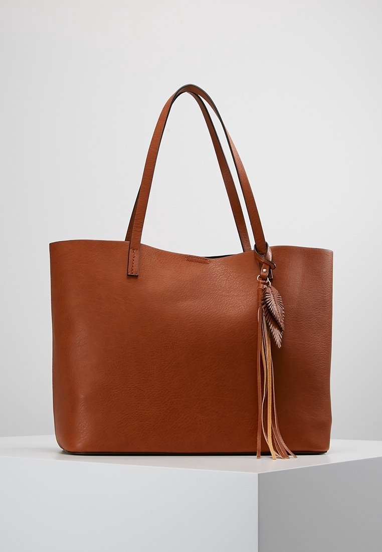 Bruine leren tote bag met twee handgrepen, voorzien van een textuur oppervlak en een decoratieve kwast met bladvormige accenten. Vierkante vorm, minimalistisch ontwerp.