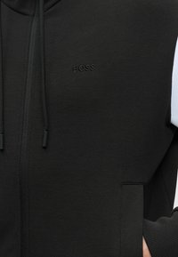 Gros plan d'un sweat à capuche noir zippé en tissu texturé, avec cordons et logo "BOSS" en relief sur la poitrine, porté par une personne avec la main dans la poche.