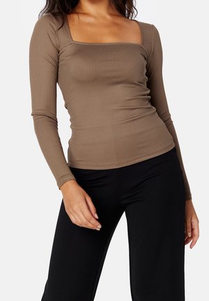 Kvinde iført en tætsiddende, ribbet, brun bluse med lange ærmer og firkantet udskæring samt sorte bukser, poserer med den ene hånd nær hoften.
