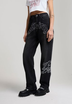 Personne portant un jean noir ample avec un texte gothique blanc et un motif de dragon, associé à des chaussures noires épaisses et un haut blanc court.