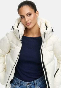 Donna con i capelli raccolti che indossa un giubbotto bianco imbottito sopra una maglia blu scuro e jeans blu, in piedi davanti a uno sfondo semplice.