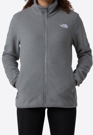 Osoba ubrana w szarą kurtkę polarową z przednim suwakiem i logo The North Face na lewej piersi, ręce w bocznych kieszeniach.