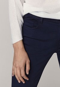 Marineblauwe broek gemaakt van een gladde stof, met een voorkantzak en een aansluitend ontwerp. Een gedeeltelijk getoonde witte textuurtop.