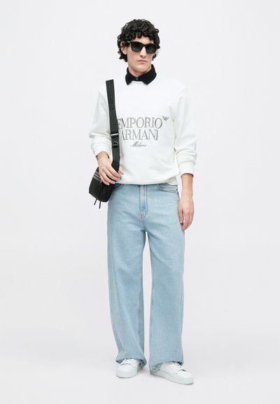 Sweatshirt branca com logo "Emporio Armani", gola escura, jeans azul claro de perna larga, mala tiracolo preta e óculos de sol pretos. Ténis brancos.