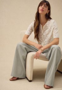 White lace short-sleeve top with a button front, paired with light gray wide-leg pants. Sitting on a cream chair with brown accents.