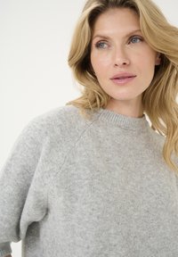Lichtgrijze trui gemaakt van zacht materiaal, met een ronde hals en raglanmouwen. De textuur lijkt glad met minimale patronen.