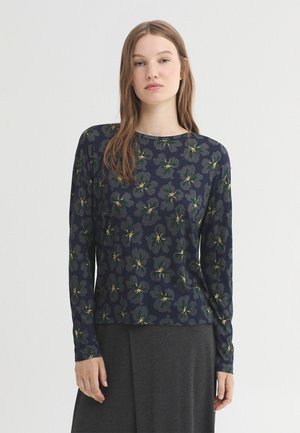 Jeune femme portant une chemise à manches longues bleu marine avec des motifs floraux verts et une jupe gris foncé, debout devant un fond blanc.