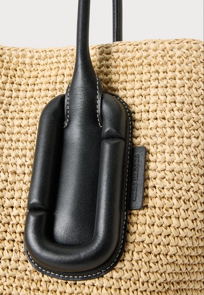 Sac beige tissé avec une grande anse en cuir noir et une étiquette avec le logo Marc Jacobs visible sur le côté.