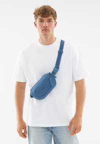 Sininen olkalaukku, jossa on tyylikäs muotoilu, pehmeä materiaali, vetoketjullinen osasto ja säädettävä hihna. Päällä valkoinen T-paita.