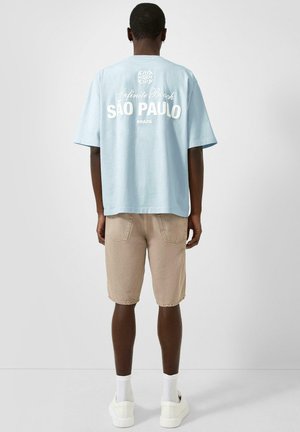 Man wearing light blue t-shirt with "Infinite Beach São Paulo Brazil" text, beige shorts, white socks, and white sneakers standing facing away.