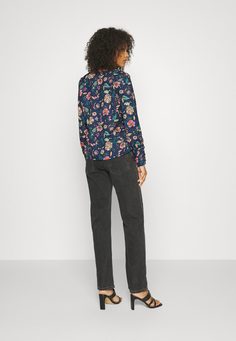 Top de mangas longas floral em azul-marinho com padrões coloridos, combinado com calças cinza-escuro e sapatos de salto preto com tiras, exibindo uma silhueta ajustada.