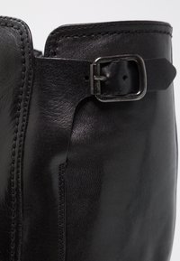 Gros plan sur une botte en cuir noir montrant des coutures cousues et une lanière avec boucle métallique près du bord supérieur.