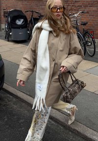 Beige oversized jas, lange witte sjaal met franjes, bloemenpatroon broek, beige hakken en een handtas van slangenprint. Ronde zonnebril.