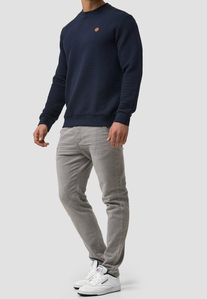 Marineblauwe trui met honingraatstructuur, ronde hals en geribbelde manchetten, gecombineerd met lichtgrijze slim-fit broek en witte sneakers.