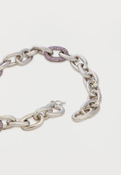 Bracelet chaîne argentée épaisse à maillons ovales, avec un seul maillon orné de pierres violettes et un fermoir, sur fond blanc.