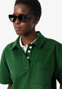 Grünes Baumwoll-Poloshirt mit weißem Kragen, vier Knöpfen, einer Brusttasche und gesticktem Logodetail. Schwarze, eckige Sonnenbrille mit gemusterten Bügeln.