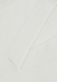 Top blanc à col en V, fabriqué en tissu doux et texturé. Lignes épurées et design minimaliste avec des couches superposées au niveau de l'encolure.