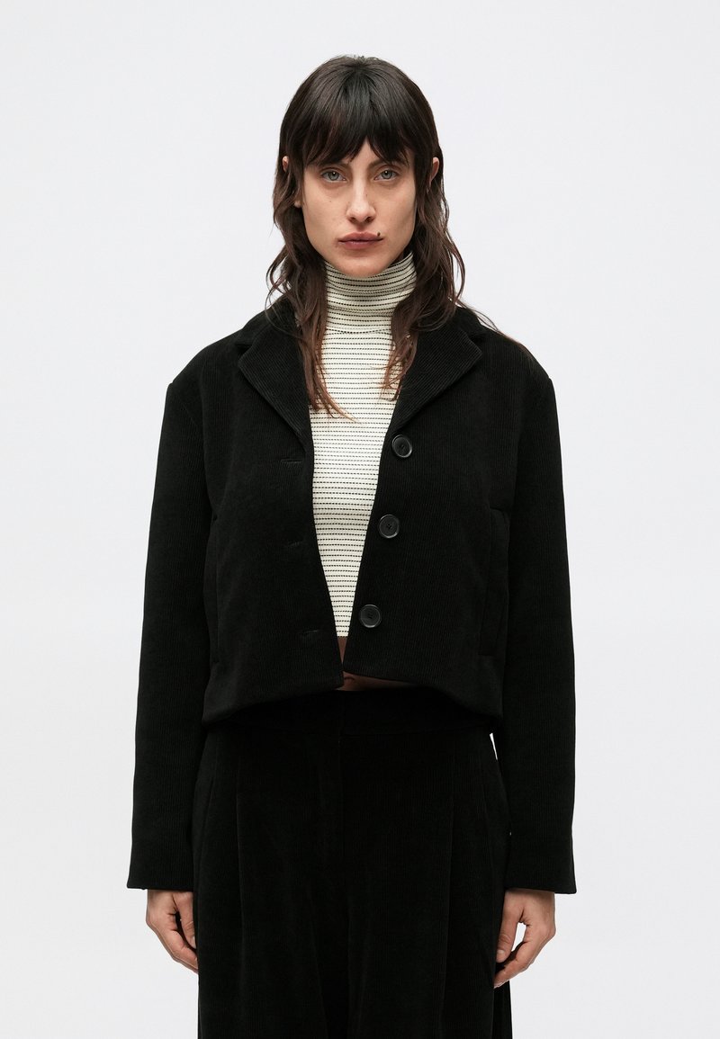 Fekete kordbársony rövidített dzseki kivágott gallérral, gombos elülső résszel és hosszú ujjakkal, világos csíkos garbó fölött viselve.