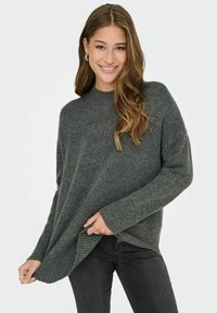 Pull en tricot gris avec un col montant, des épaules tombantes et des poignets côtelés. Le tissu a une texture douce et une coupe décontractée.