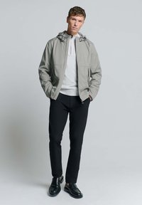 Lichtgrijze jas met capuchon, ritssluiting aan de voorkant, twee zijzakken, gedragen over een wit shirt, gecombineerd met zwarte broek en zwarte instapschoenen.