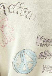 Tessuto bianco con disegni stampati: orsetto rosa, "Action" in nero e simbolo di pace blu. Dettagli tattili e stile morbido e casual.