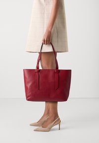 Bolsa tote em couro vermelho com acabamento texturizado, duas alças e correias longas. Combinada com um vestido plaid até ao joelho e sapatos de salto alto bege.