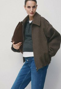 Kobieta ubrana w brązową, kratkowaną kurtkę na ciemnoszarym swetrze i niebieskich dżinsach, trzymająca dużą brązową torebkę kopertówkę na jednolitym tle.