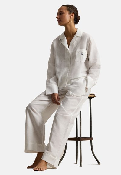 Vrouw zit op een kruk, op blote voeten, en draagt een los witte blouse met knopen en bijpassende broek met wijde pijpen en geborduurde logo's.