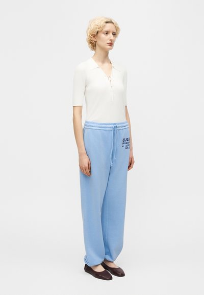 Νεαρός με κοντά σγουρά ξανθά μαλλιά που φοράει λευκό κοντομάνικο μπλουζάκι, ανοιχτόχρωμο μπλε άνετο παντελόνι φόρμας με κορδόνι και σκούρα επίπεδα παπούτσια.