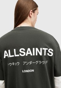 Camiseta de algodón verde oscuro con grandes letras blancas "ALLSAINTS" y otros idiomas. Cuello redondo simple, corte holgado y mangas cortas.