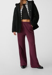 Schwarze Kunstfelljacke mit Kapuze über einem schwarzen Croptop, kombiniert mit lockeren mauroroten Hosen mit seitlichen Streifen und schwarzen flachen Schuhen.
