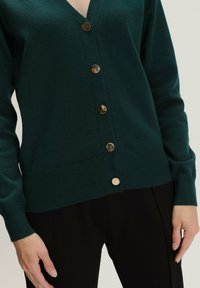 Dunkelgrüner Cardigan aus weichem Stoff mit V-Ausschnitt, versehen mit sechs goldfarbenen Knöpfen sowie gerippten Bündchen und Saum.