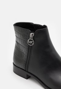 Botín negro hasta el tobillo con parte delantera de cuero liso, parte trasera con patrón de cocodrilo texturizado, cremallera lateral y tirador redondo de metal plateado con logo.