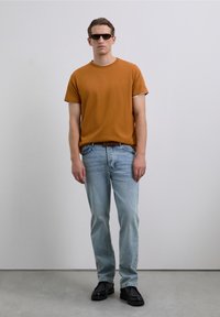 Man wearing orange t-shirt, light blue jeans, black shoes, and sunglasses standing against plain gray background.