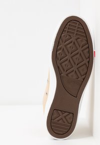 Tan canvas sneaker with white rubber sole, featuring textured brown outsole and silver eyelets. Logo visible on the side.