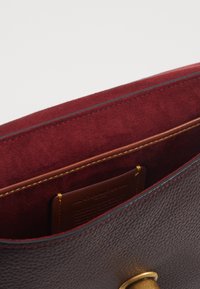 Nahkainen käsilaukku, jonka sisäpuoli on tummaa burgundinsinistä mokkanahkaa, kullanväriset metalliosat ja teksturoitu ulkopuoli. Sisältää avoimen taskun ja kirjailtuja koristeita.