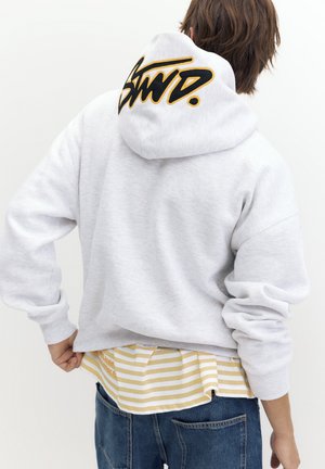 Personne portant un sweat à capuche gris clair avec un logo "SWD." noir et jaune sur la capuche, une chemise rayée jaune et un jean bleu, vue de dos.