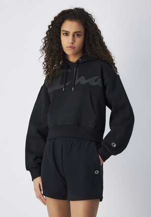 Felpa nera corta con tasca frontale, maniche oversize e dettagli con logo. Abbinata a pantaloni corti neri con cintura elastica.