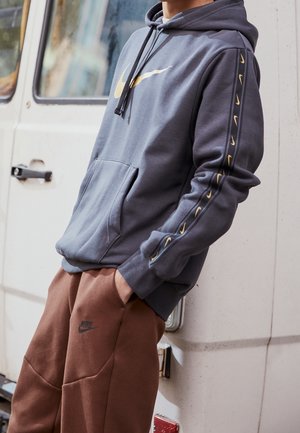 Persona vistiendo una sudadera gris de Nike con los logos dorados del swoosh en el pecho y la manga, y pantalones de chándal marrones de Nike, apoyado contra un vehículo blanco.