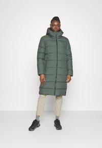 Donkergroene pufferjas, tot op de knie, met horizontale stiknaden, hoge kraag en een ritssluiting aan de voorkant. Gecombineerd met beidgekleurige broek en zwarte schoenen.