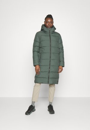 Doudoune de couleur vert foncé, longueur genoux, avec des coutures horizontales, un col montant et une fermeture éclair à l'avant. Associée à un pantalon beige et des chaussures noires.