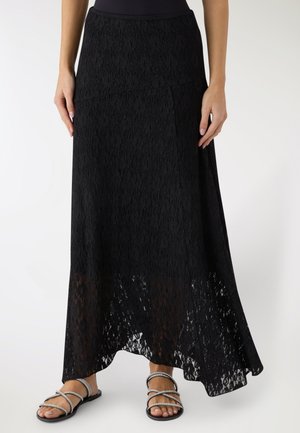 Gonna lunga in pizzo nero con orlo asimmetrico, indossata con sandali piatti argento con cinturini, mostrata dalla vita ai piedi su sfondo bianco.