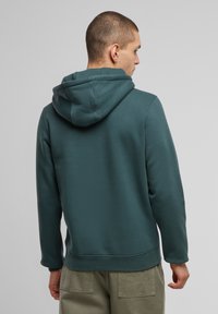 Mikina s kapucňou v tmavom teal odtieni, vyrobená z mäkkej tkaniny. Má voľný strih a rebrované manžety. Jednoduchý dizajn bez viditeľných log. 