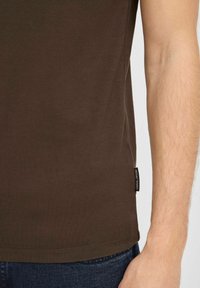 T-shirt en coton marron avec col rond et manches courtes, doté d'une étiquette sur le côté près de l'ourlet. Texture lisse et coupe ajustée.