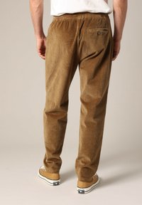 Bruine corduroy broek met een rechte pijp, voorzien van een achterzak en een elastische tailleband. Gedragen met tan schoenen, tegen een neutrale achtergrond.