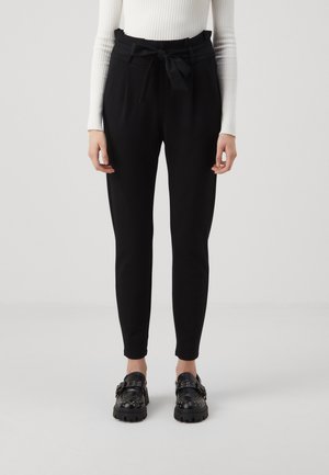 Schwarze, schmal zulaufende Hosen mit hoher Taille, einem Stoffgürtel mit Schleife und glatter Textur; kombiniert mit glänzenden schwarzen Slippern.