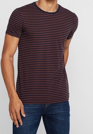 Man wearing navy blue and brown horizontal striped short-sleeve t-shirt and dark blue jeans, standing with one hand in pocket.