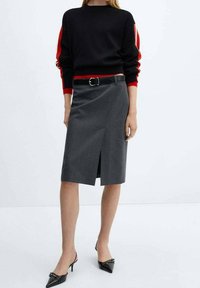 Harmaa muotosaumattu kynähame, jossa on etupuolella halkio, yhdistettynä mustaan neuleeseen, jossa on punaisia raitoja. Musta vyö ja kärkitossut täydentävät asua.